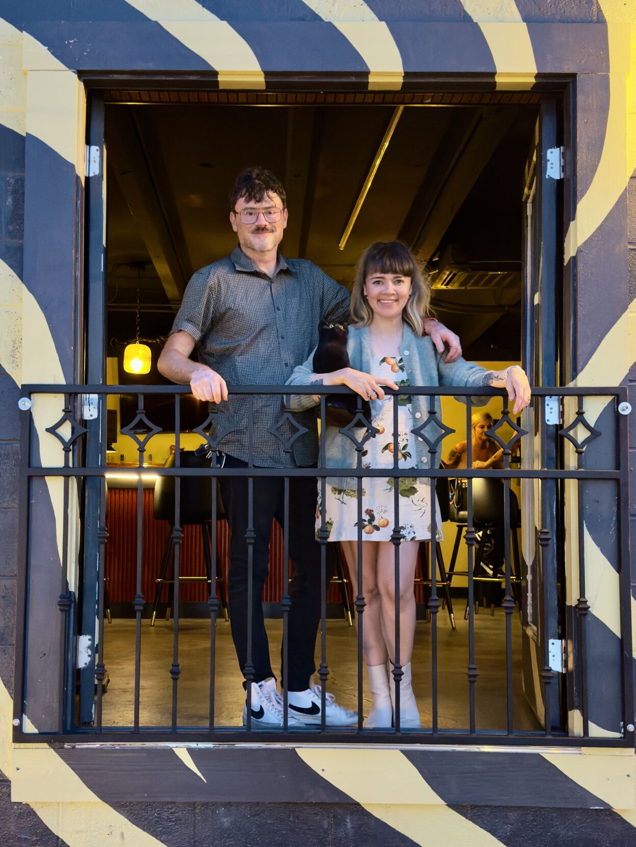 A couple stands on a balcony holding a black cat with a bar visible behind them